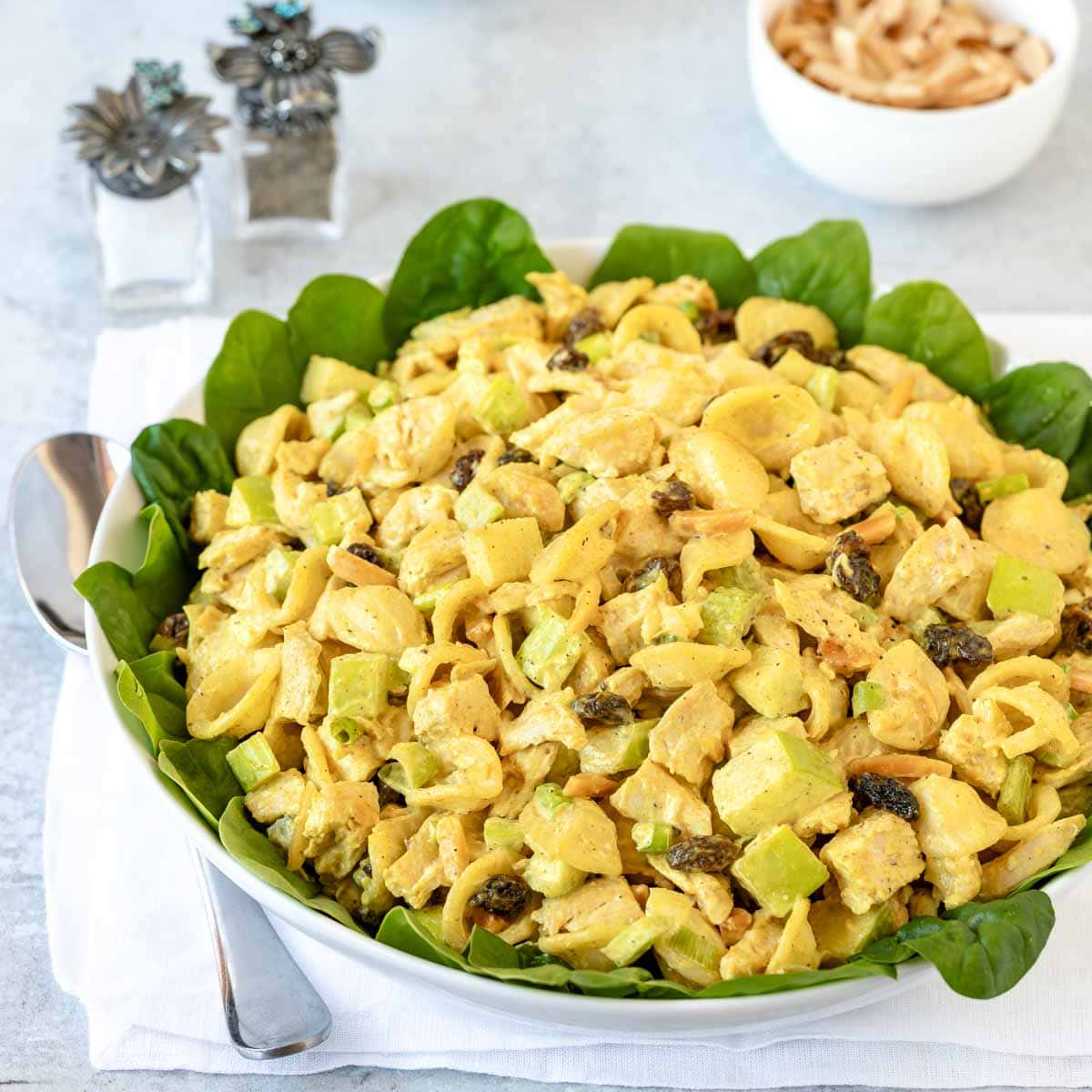 bowl lined with spinach leaves holding Curry Chicken Pasta Salad with salt, pepper, and a small bowl of almonds