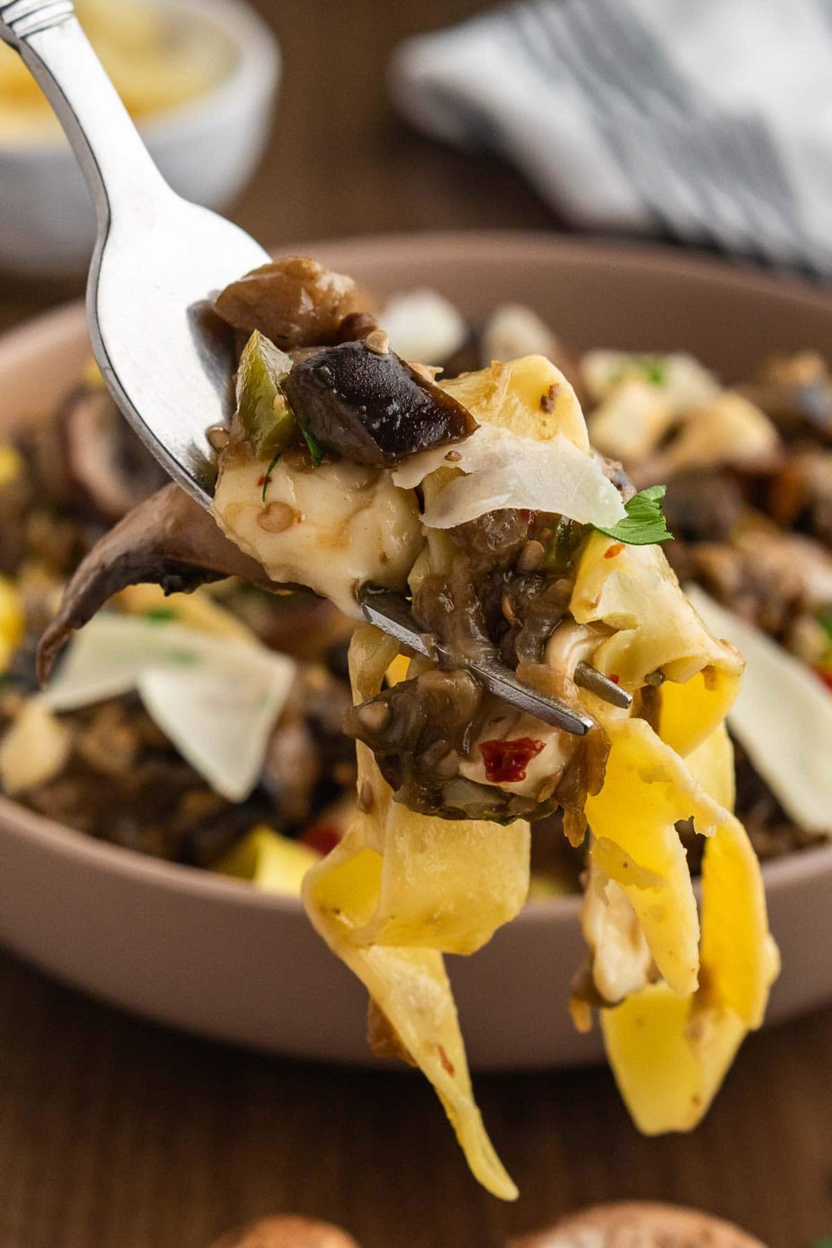 A forkful of eggplant and mushroom pasta held up over a bowl of more.
