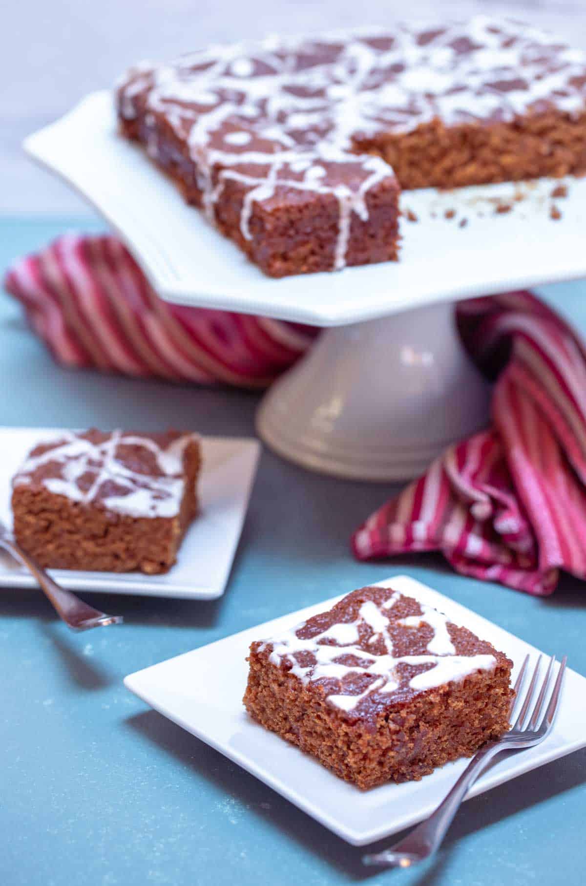 Two square plates holding slices of Sticky Ginger Cake, with cake in the background.