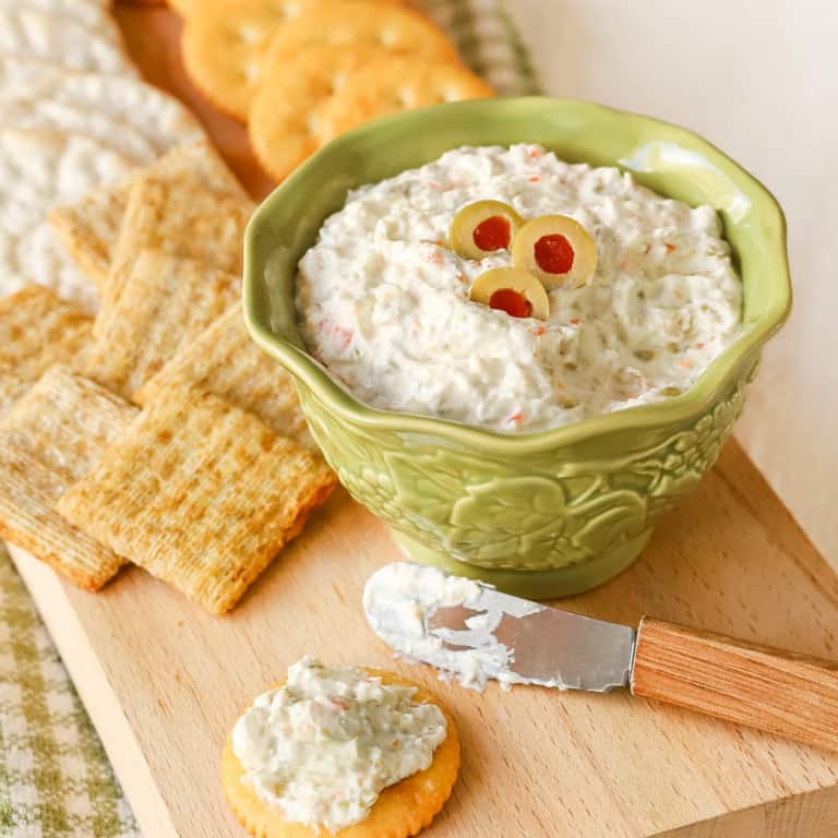 Small bowl holding Green Olive Cheese Spread, with some on a cracker in front and more crackers in back