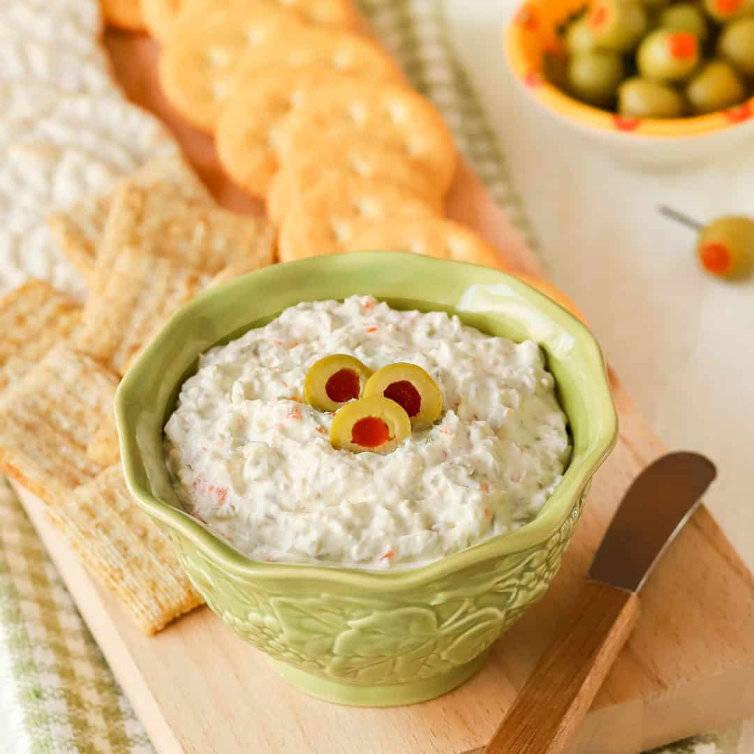 Small bowl holding Green Olive Cheese Spread, with some on a cracker in front and more crackers in back