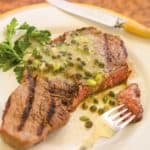 White plate holding one grilled ribeye steak, with a bite on a fork