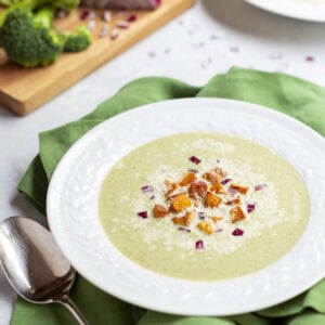 white bowl holding one serving of Healthy Broccoli Soup with potato croutons, red onion and Parmesan cheese garnishes. broccoli florets and partially chopped red onion in the background