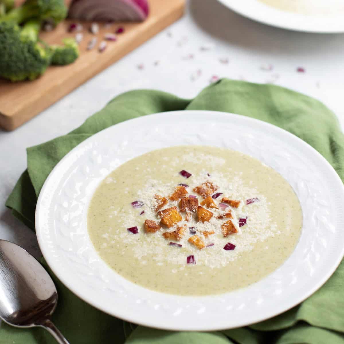 White bowl with Healthy Broccoli Soup topped with Potato Croutons, red onion and Parmesan