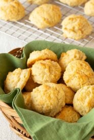 Wicker basket lined with a green napkin and filled with Easy Drop Biscuits, with more biscuits on a cooling rack in the back