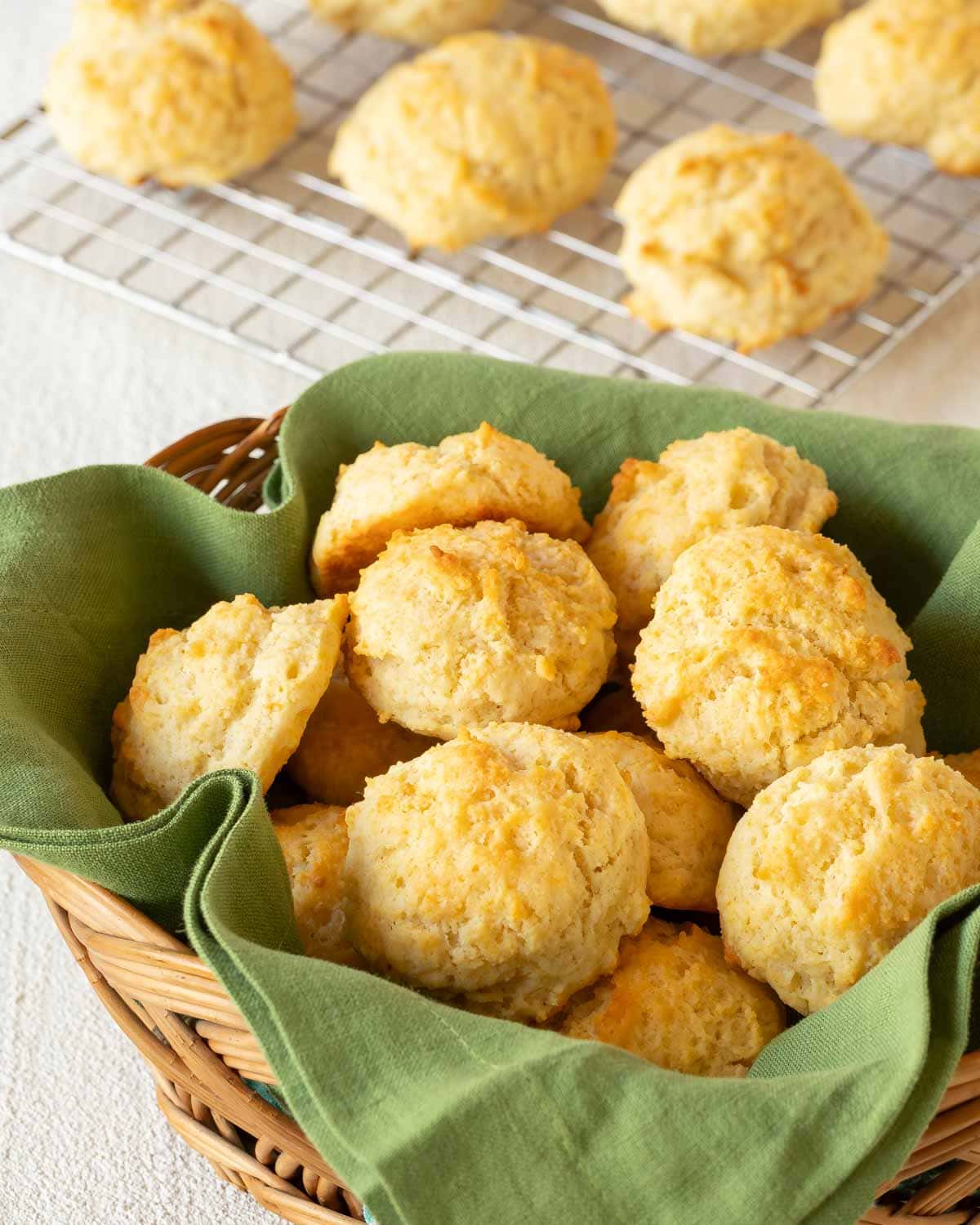 Wicker basket lined with a green napkin and filled with Easy Drop Biscuits, with more biscuits on a cooling rack in the back