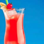 Close up of tall glass filled with red hurricane cocktail garnished with orange and lime slices and a cherry.