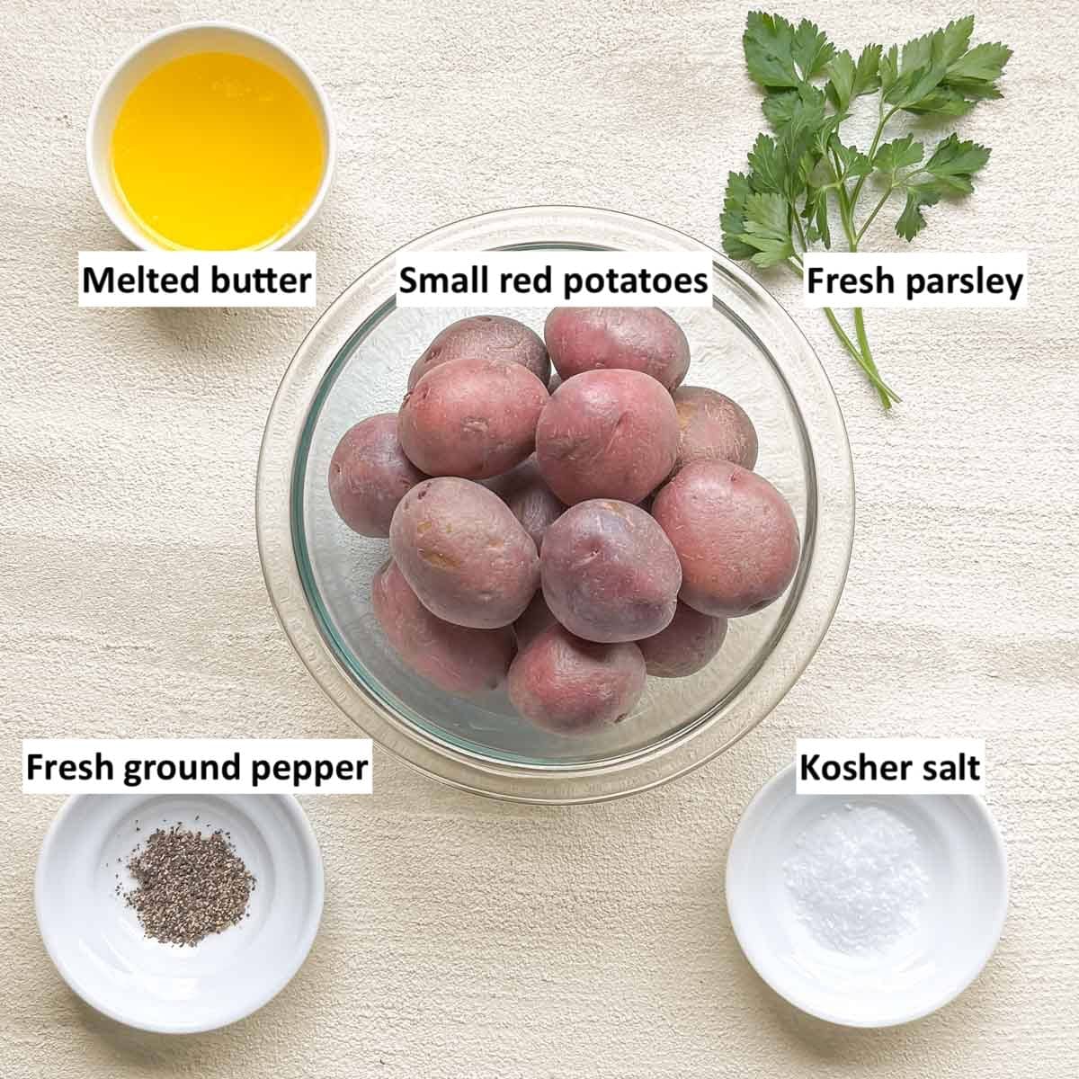 bowl holding small red potatoes, surrounded by other recipe ingredients, including melted butter, fresh parsley, kosher salt, and ground pepper.