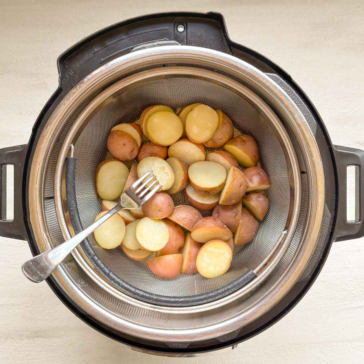 Steamed halved small red potatoes inside Instant Pot, in steamer basket, with fork poking one to show texture when done.