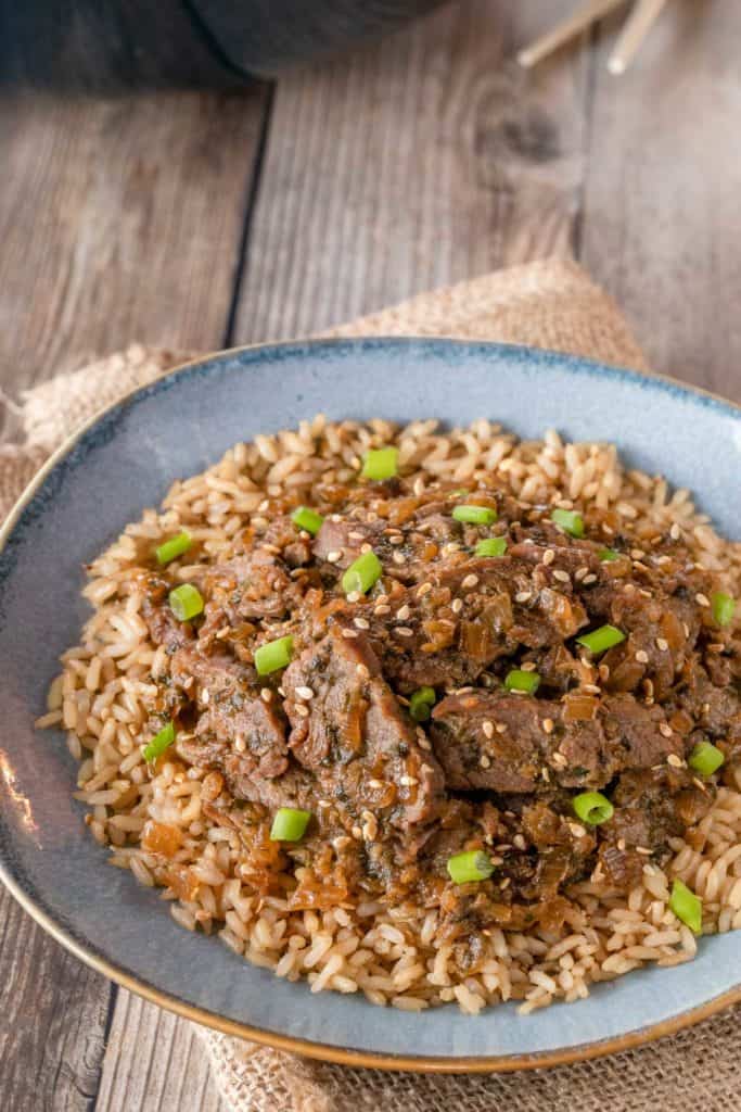 Blue plate holding Korean BBQ Beef over brown rice. Plate is sitting on a brown napkin