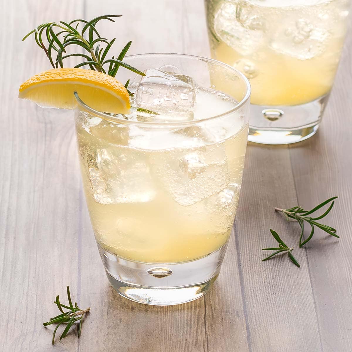 close up of two clear cocktail glasses holding Lemon Ginger Mocktails, garnished with lemon wedge and rosemary sprig