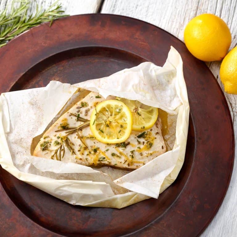 Swordfish fillet topped with lemon slices, lemon zest and chopped rosemary in a parhment paper packge.