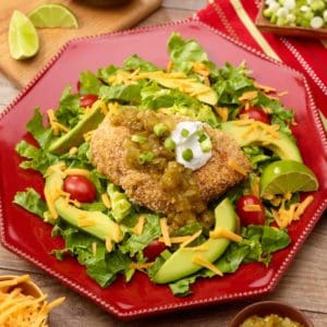 Red plate holding mexican baked chicken sitting on mixed greens, surrounded by slicd avocado and cherry tomatoes, and topped with salsa verde and sour cream.