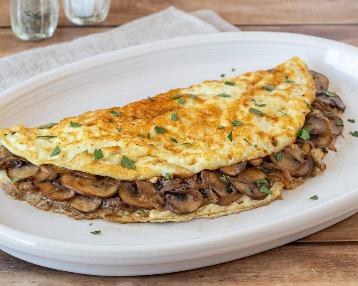 Mushroom omelet with sautéed onions and parsley on an oval white plate.