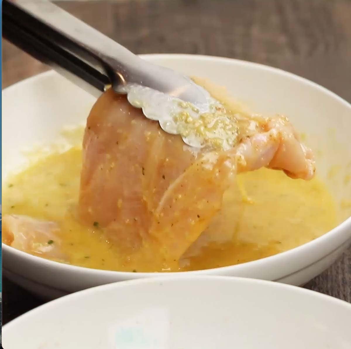 Tongs holding a chicken breast over the bowl of egg mixture. 