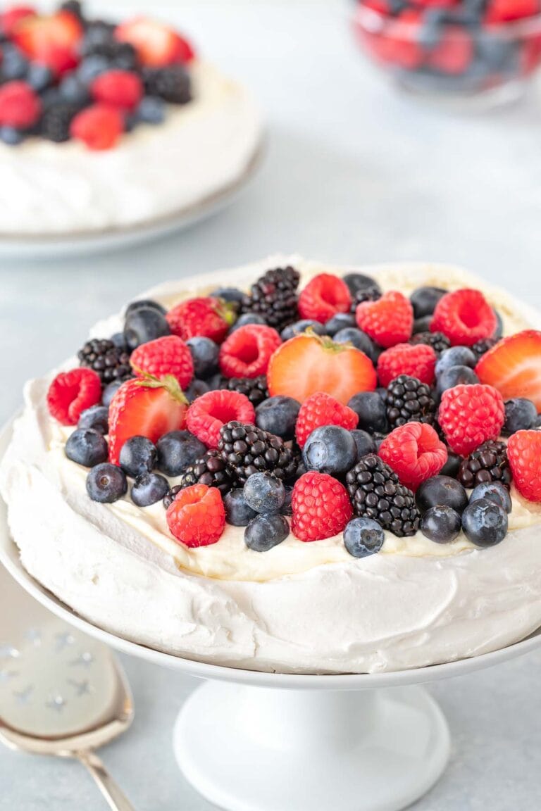 One Pavlova on a cake stand, with a second one and a bowl of berries in the background