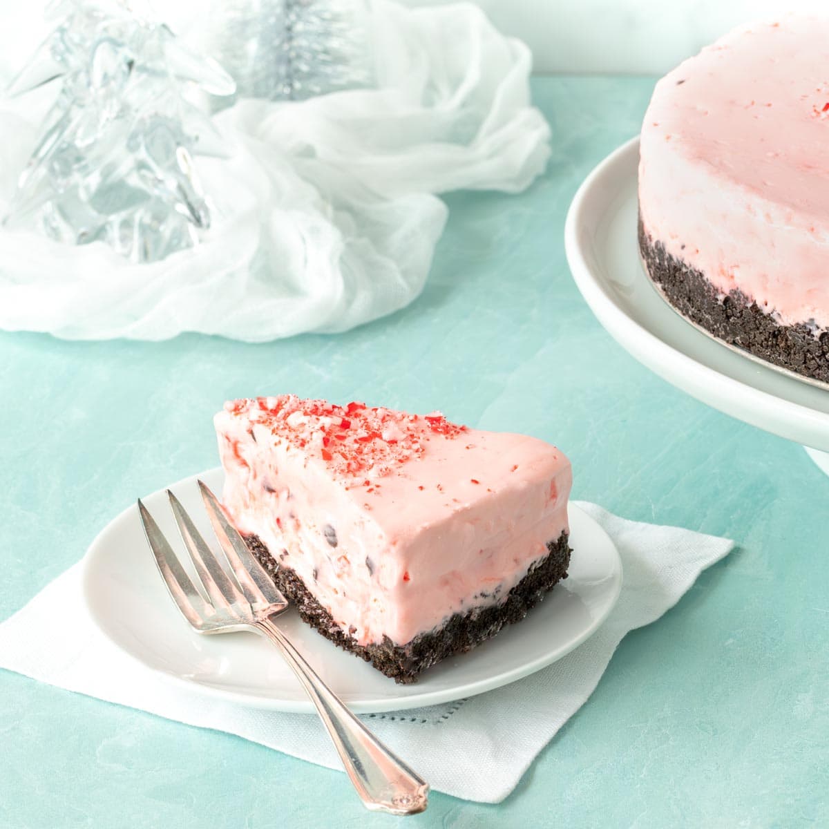 Slice of Frozen Peppermint Chocolate Cheesecake on a white plate, with the rest of the cake in the background alongside glass trees