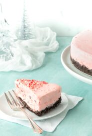 Slice of Frozen Peppermint Chocolate Cheesecake on a white plate, with the rest of the cake in the background alongside glass trees