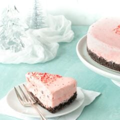 Slice of Frozen Peppermint Chocolate Cheesecake on a white plate, with the rest of the cake in the background alongside glass trees