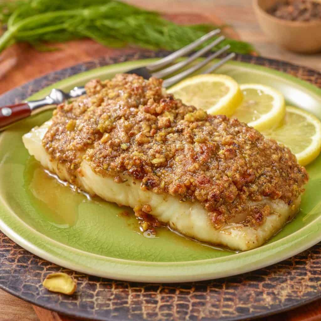 A single serving of Pistachio Crusted Fish, sitting on a light green plate that's on a cutting board. 