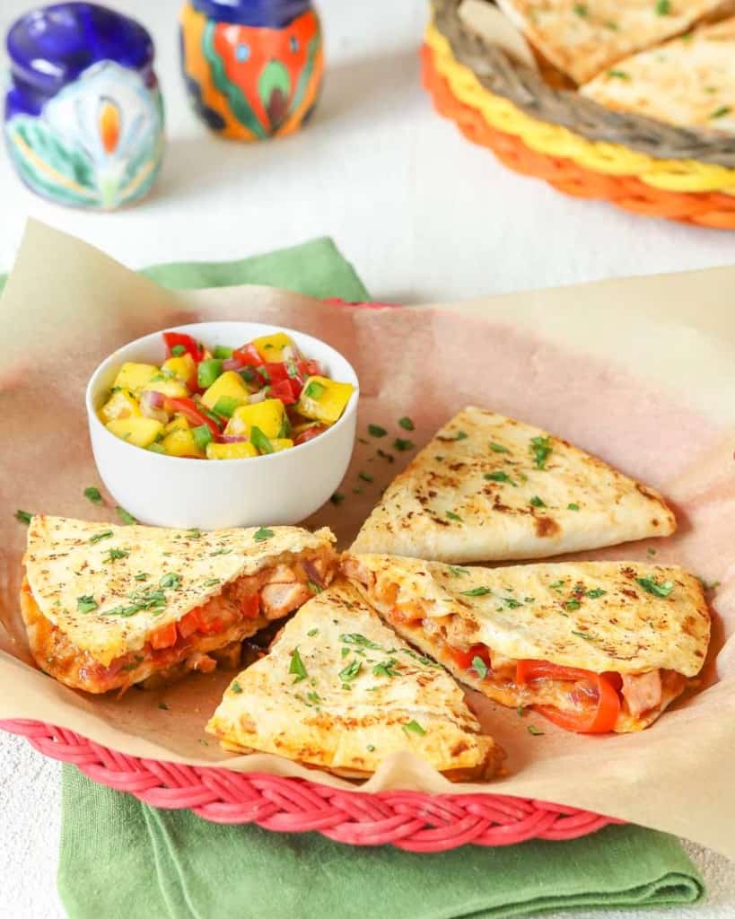 red wicker plate holding 4 slices (1 portion) of Leftover Pork Quesadillas with a small bowl of Tomato Mango Salsa alongside