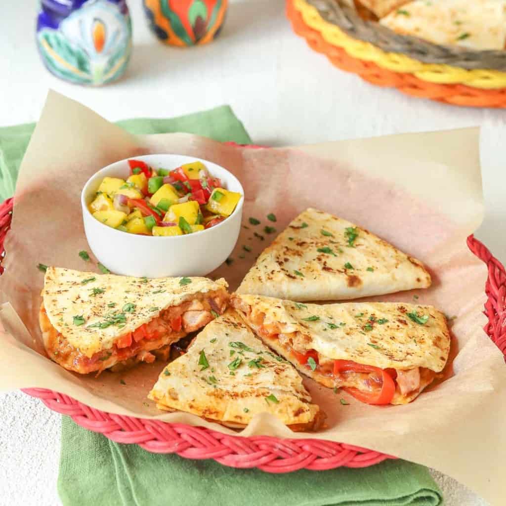 red wicker plate holding 4 slices (1 portion) of Leftover Pork Quesadillas with a small bowl of Tomato Mango Salsa alongside