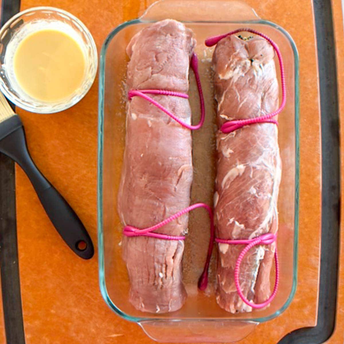 Two stuffed, rolled and secured pork tenderloins in baking dish with basting sauce on the side