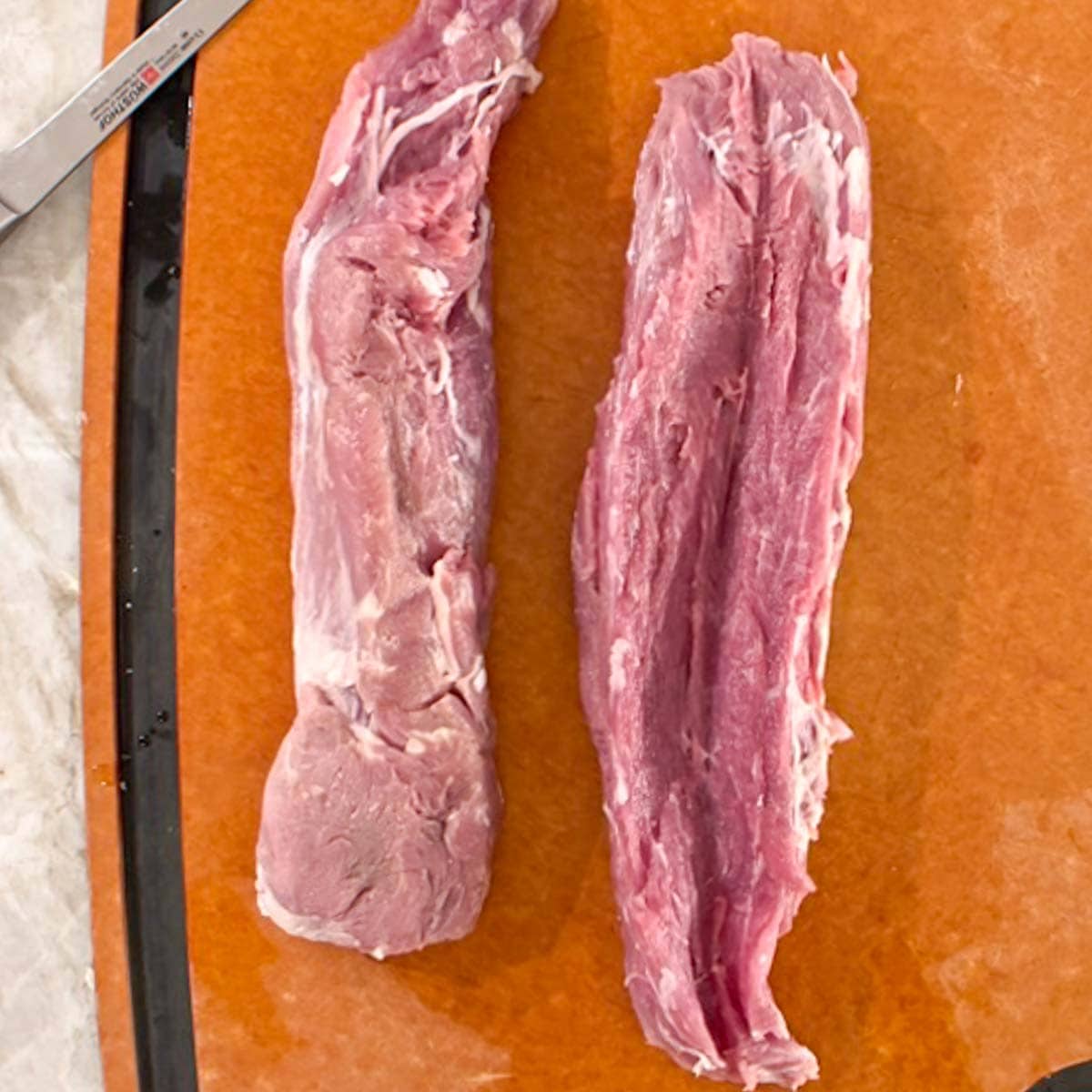 One whole raw pork tenderloin next to one that has been cut vertically, the first step in butterflying