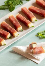 white platter holding colorful prosciutto wrapped mozzarella sticks with one stick cut in half to show inside, resting on a napkin.
