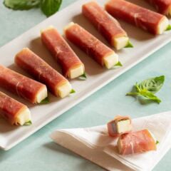 white platter holding colorful prosciutto wrapped mozzarella sticks with one stick cut in half to show inside, resting on a napkin.