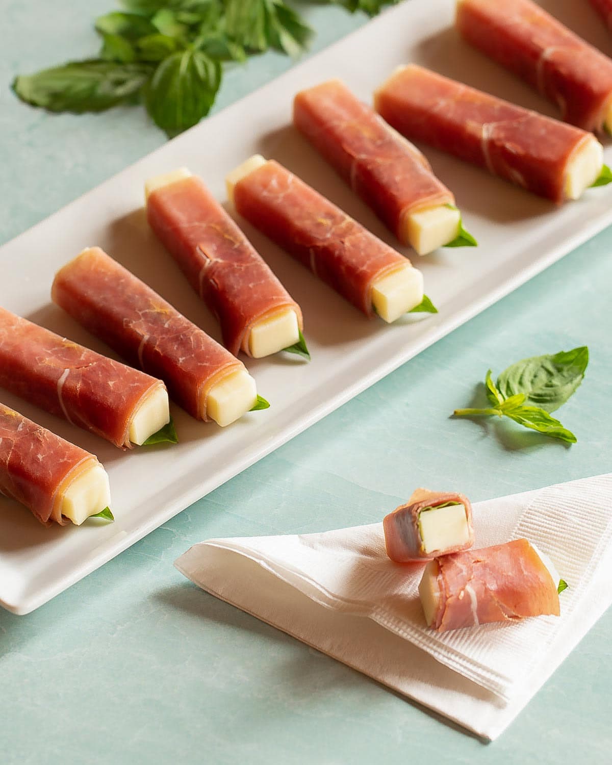 white platter holding colorful prosciutto wrapped mozzarella sticks with one stick cut in half to show inside, resting on a napkin.