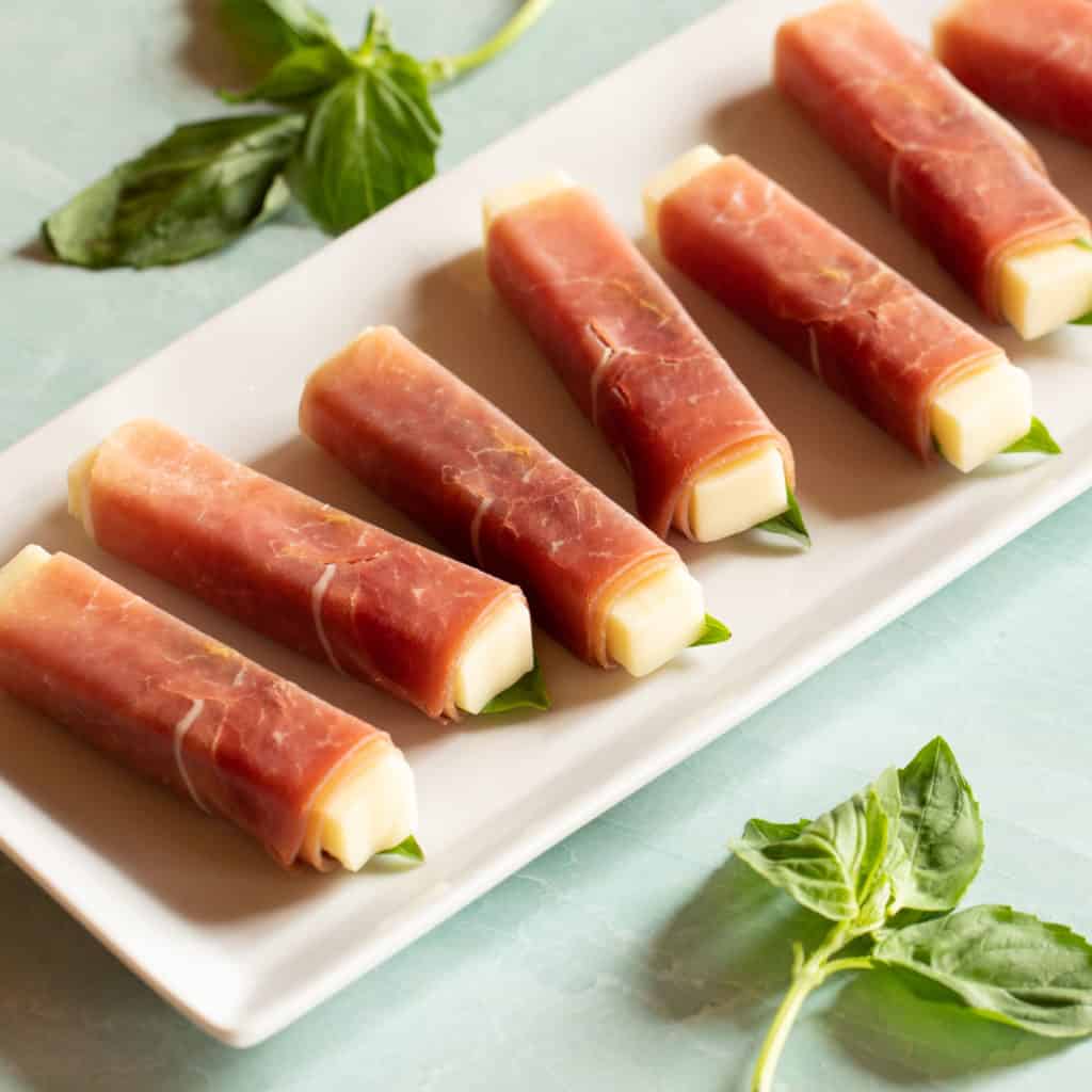 Narrow rectangular white platter filled with Prosciutto-Wrapped Smoked Mozzarella Sticks