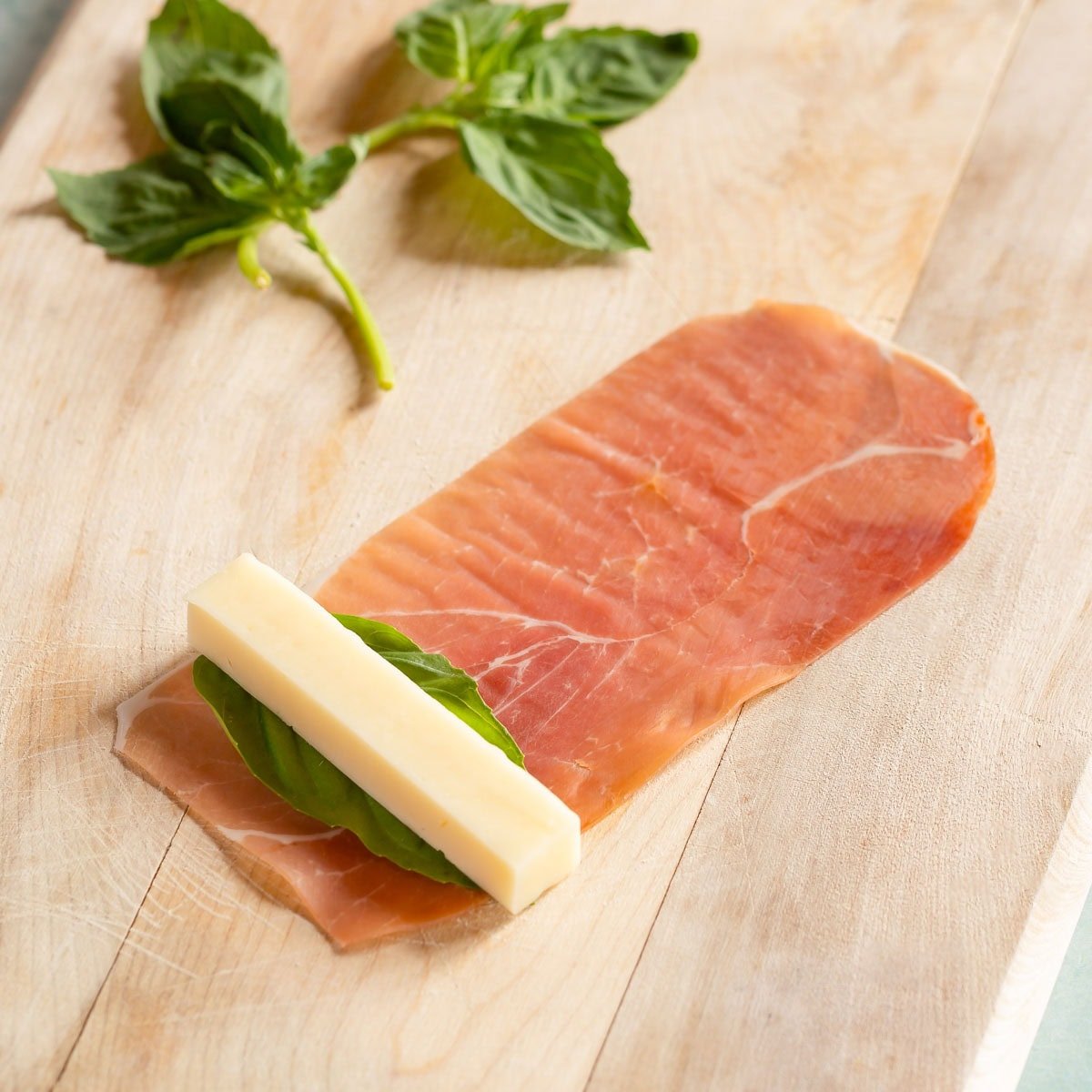 One prosciutto slice with one basil leaf and one mozzarella stick at one end, ready to be rolled.