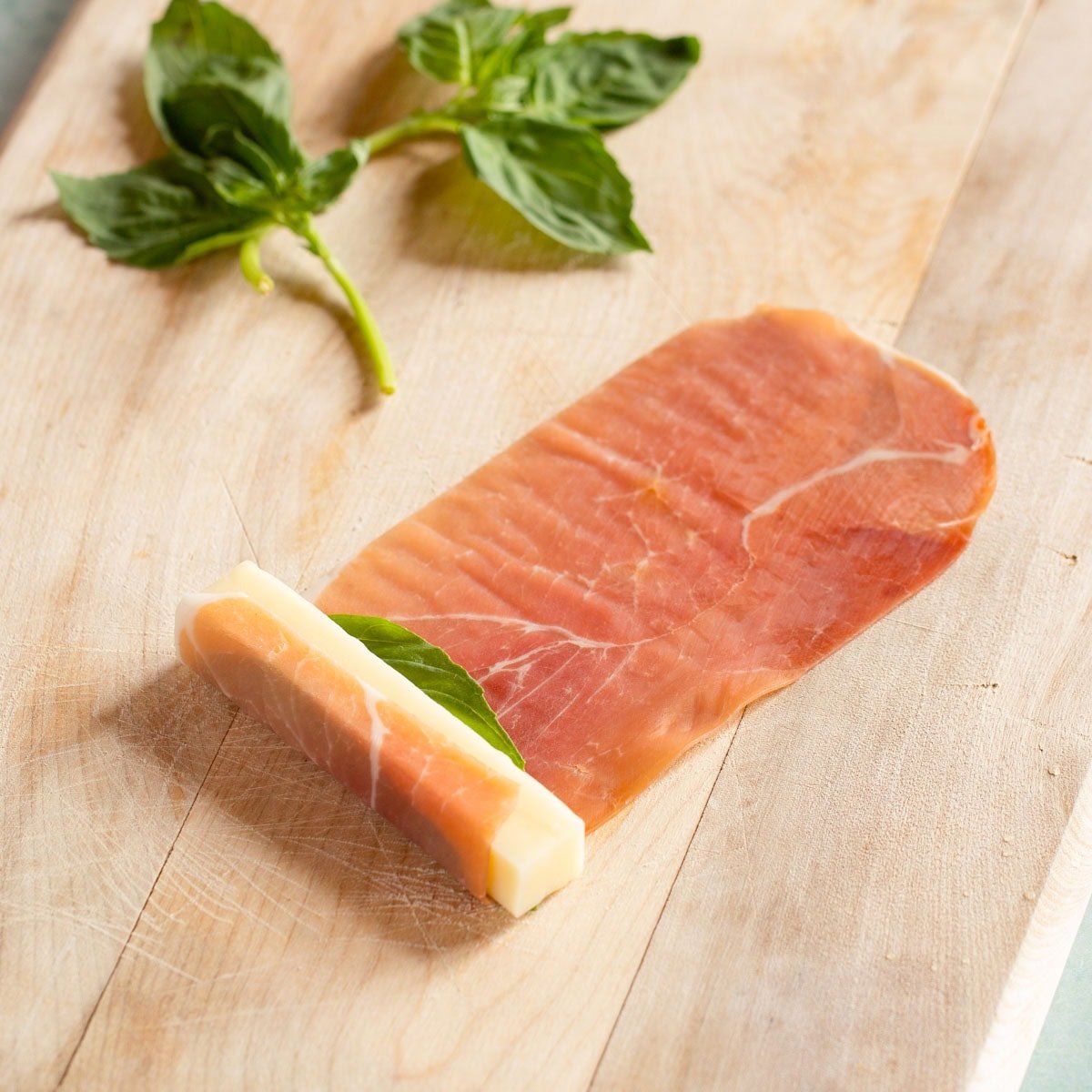 One prosciutto slice with one basil leaf and one mozzarella stick at one end, with prosciutto wrapped over the top.