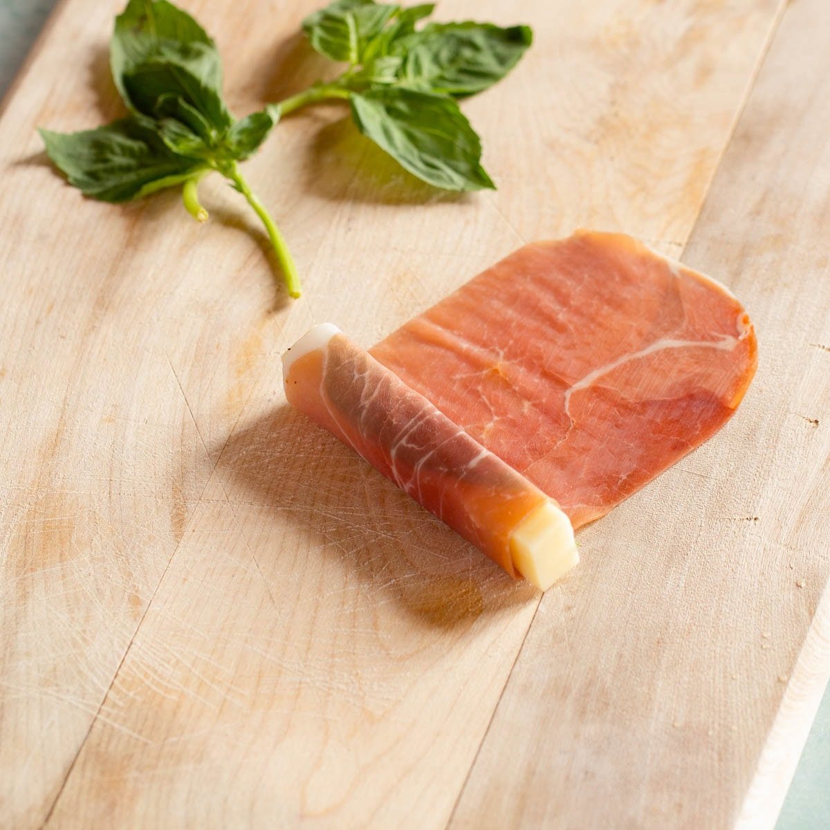 One prosciutto slice with one basil leaf and one mozzarella stick at one end, prosciutto rolled over the top 2 times.