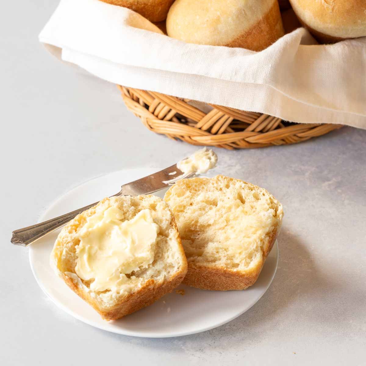 quick dinner roll split open on a plate with butter, wtih basket of more rolls in the background