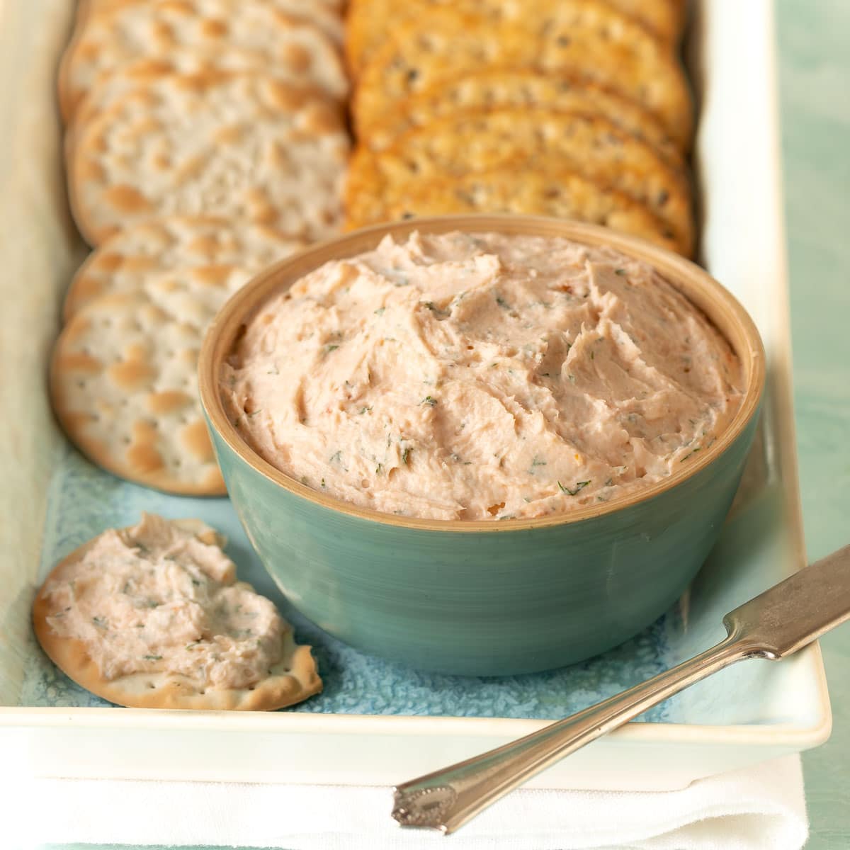 Close up shot of Smoked Salmon Mousse in a blue bowl, with a cracker with mousse on it to the side and more crackers in the back