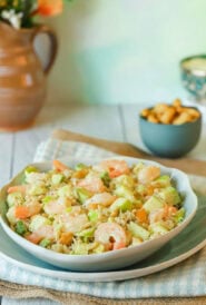Chopped Shrimp Salad with Rice and Avocado mounded on top of a blue plate, with cashews and a pitcher if flowers in the background