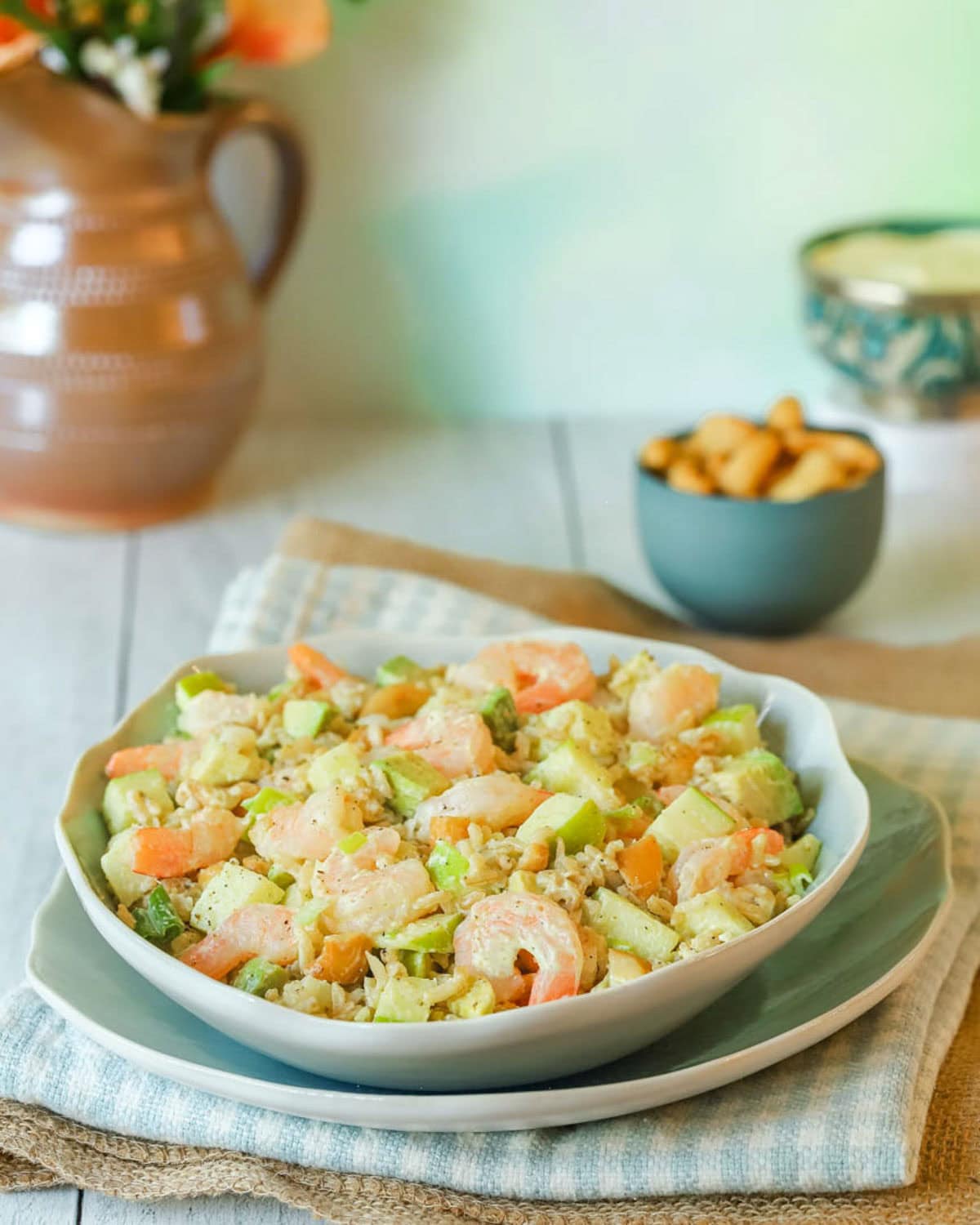 Chopped Shrimp Salad with Rice and Avocado mounded on top of a blue plate, with cashews and a pitcher if flowers in the background.