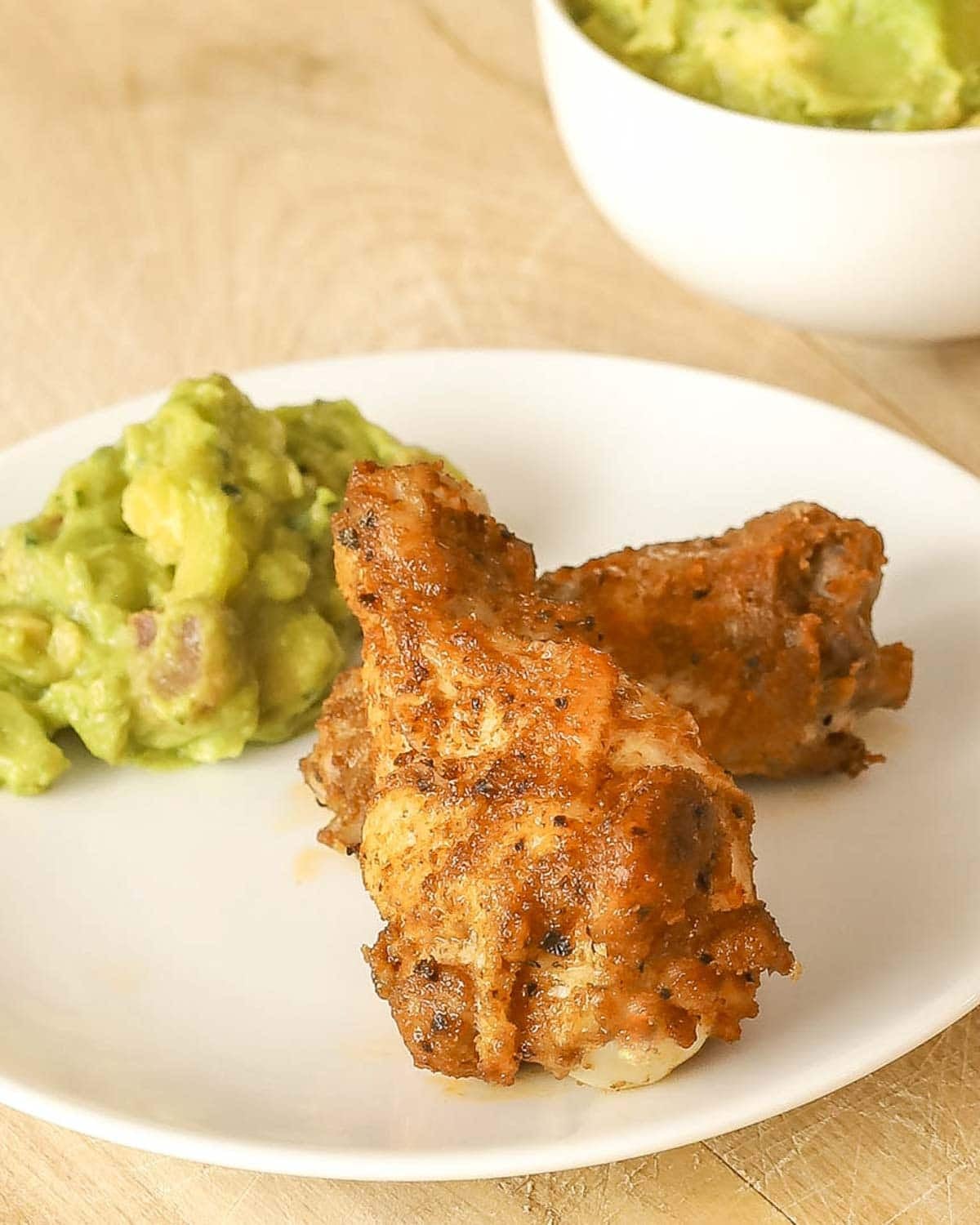 Small white plate holding 2 spicy, juicy chicken wings with a dollop of chunky guacamole.
