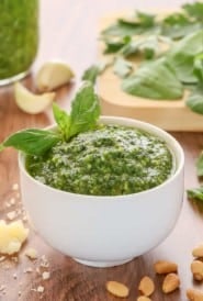 white bowl holding spinachh basil pesto, garnished with a basil leaf and with spinach and more pesto in the background