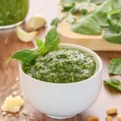 white bowl holding spinachh basil pesto, garnished with a basil leaf and with spinach and more pesto in the background