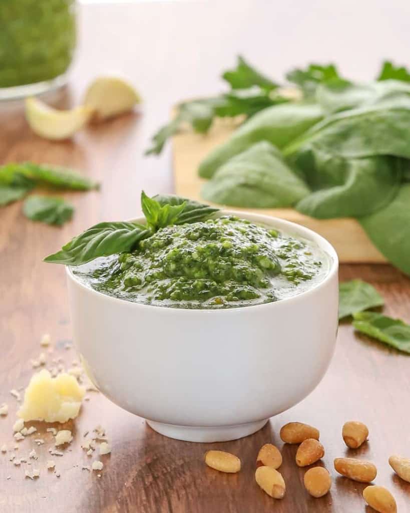 white bowl holding spinachh basil pesto, garnished with a basil leaf and with spinach and more pesto in the background