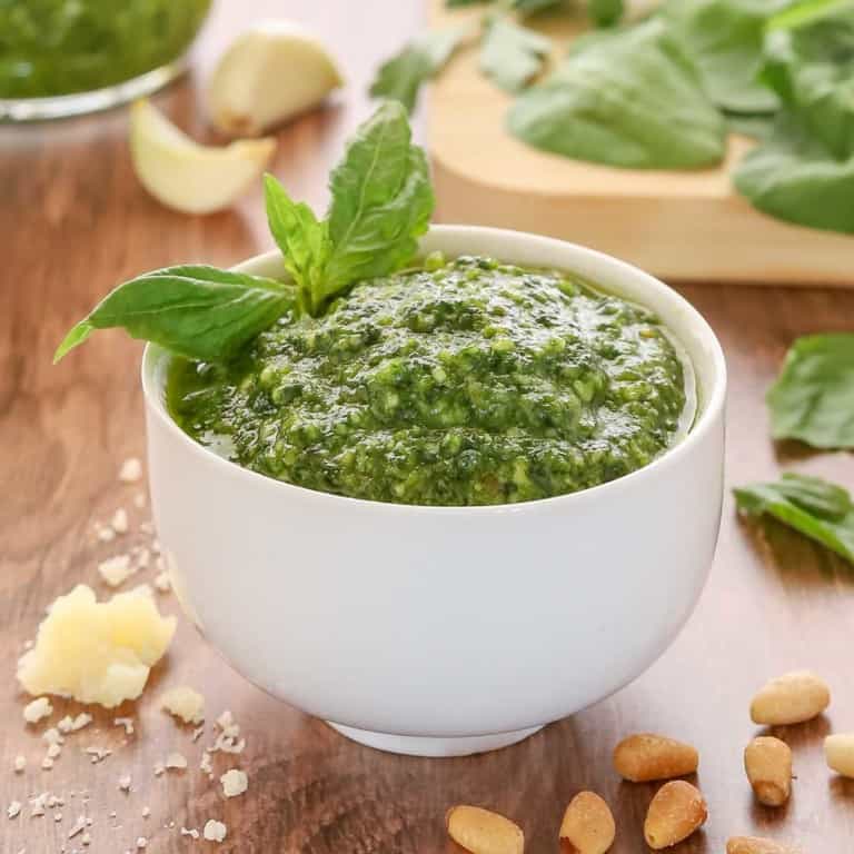 white bowl holding spinachh basil pesto, garnished with a basil leaf and with spinach and more pesto in the background