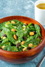 wooden bowl holding a serving of spinach salad with raisins and peanuts and dressing on the side