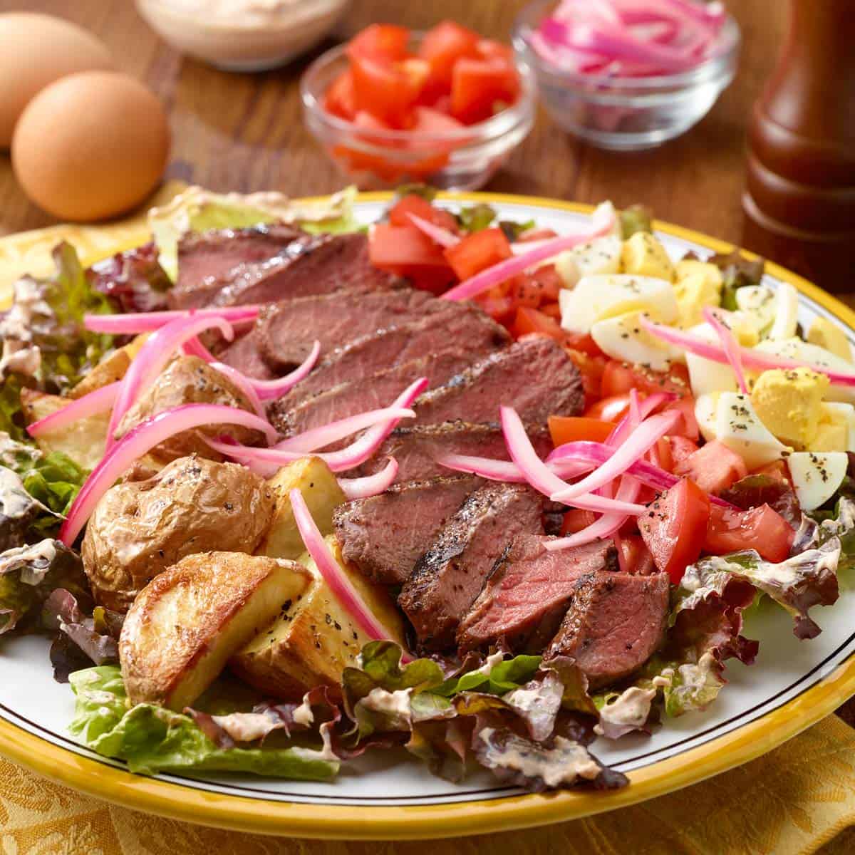 Steak and potato salad plated on a colorful plate with ingredients in rows on top of the bed of lettuce.