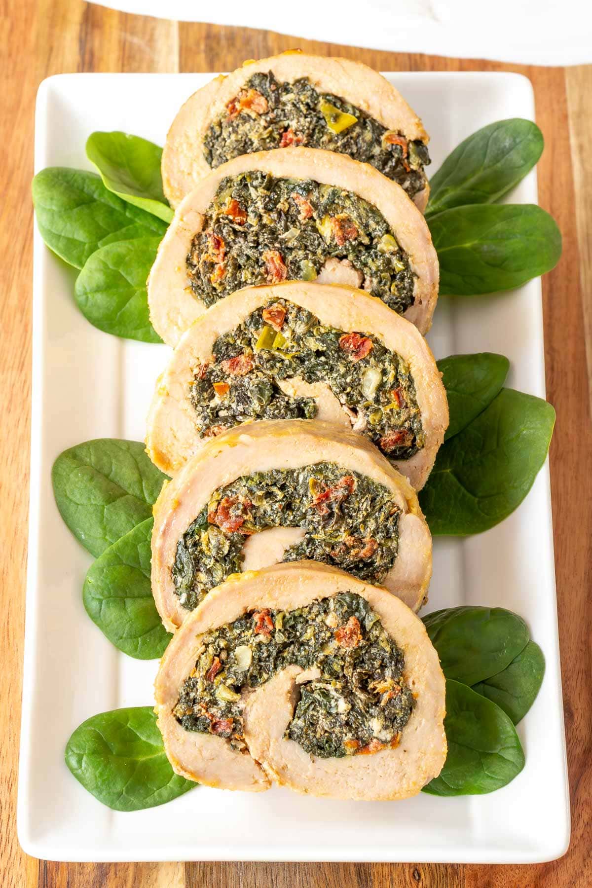 Five slices of stuffed pork tenderloin on a white platter, garnished with baby spinach on the side