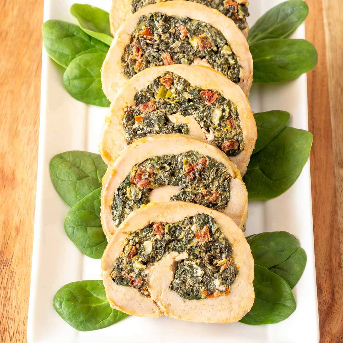 Four slices of stuffed pork tenderloin on a white platter, garnished with baby spinach on the side