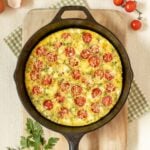 cast iron skillet holding a Tomato Frittata with Feta, on top of a cutting board and surrounded by cherry tomatoes, fresh parsley and dill, feta in a small bowl, and egg shells