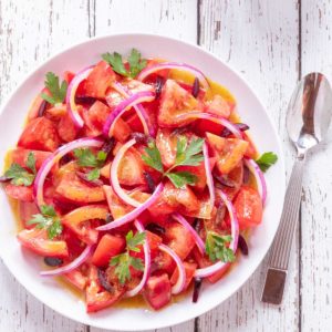 White bowl holding chopped tomatoes and slivered black olives tossed in cumin dressing and topped with sliced red onion and fresh parsley leaves.